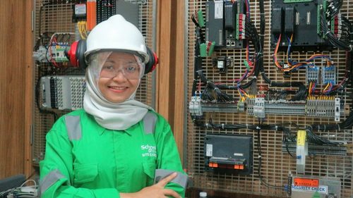 Maulidya Falah pictured, wearing a hard hat and ear-protectors standing in front of a wall of electronic cables and devices