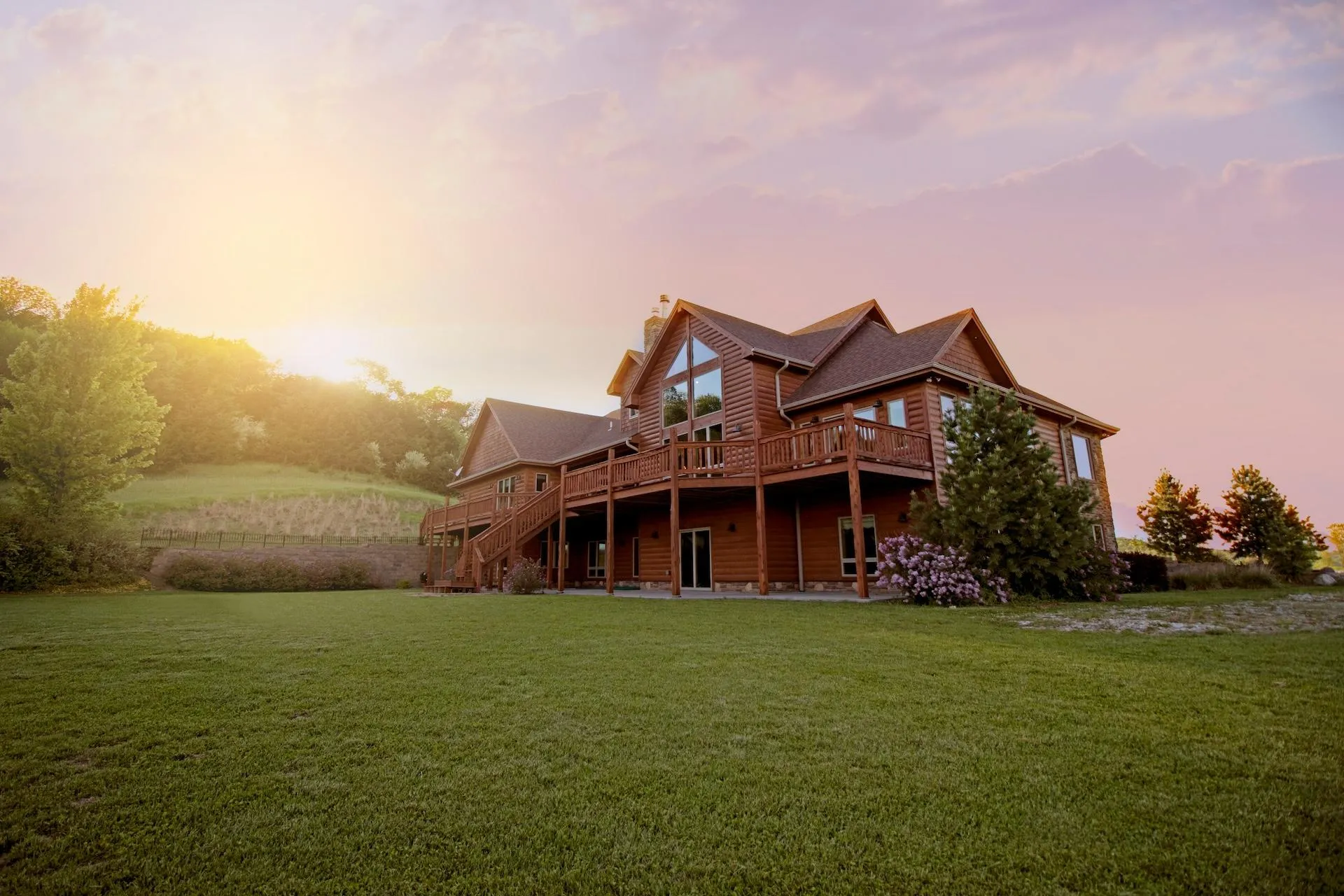 A two-story wooden house with multiple balconies and a spacious lawn, set against a sunset sky.