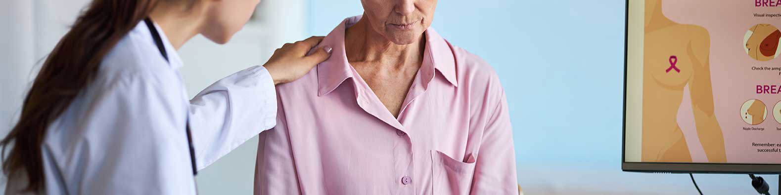 Eine Ärztin legt beruhigend ihre Hand auf die Schulter einer älteren Frau in rosa Bluse, während auf einem Bildschirm daneben eine Grafik zur Brustuntersuchung zu sehen ist.