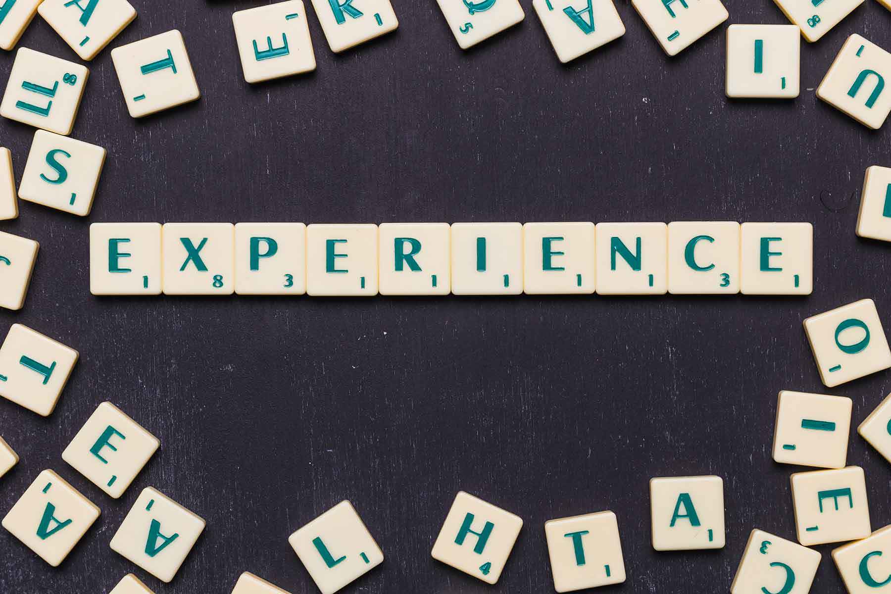 Word “EXPERIENCE” spelled out with Scrabble letter tiles arranged in a straight line on a dark background, surrounded by scattered tiles with random letters.
