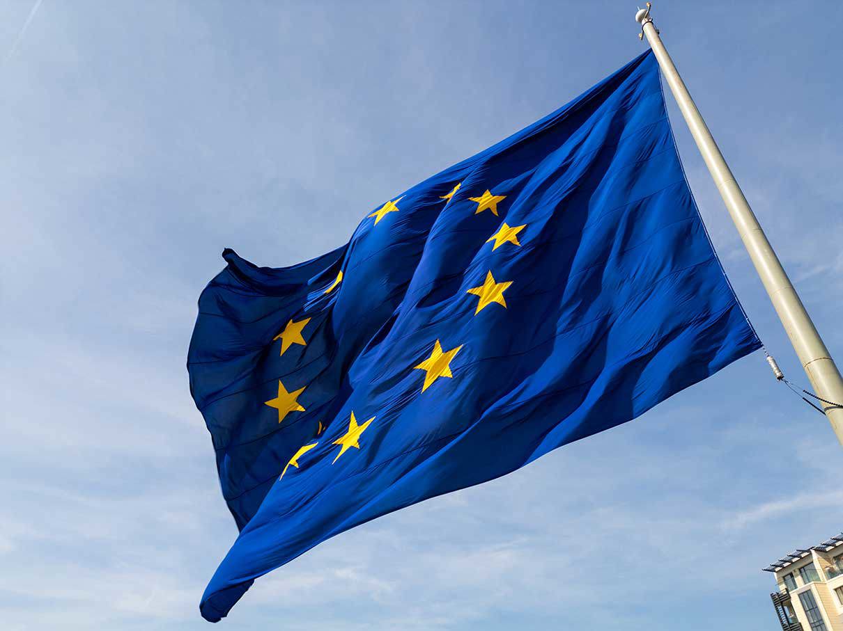 Die Flagge der Europäischen Union mit zwölf goldenen Sternen auf blauem Hintergrund weht an einem Fahnenmast vor einem hellen Himmel.