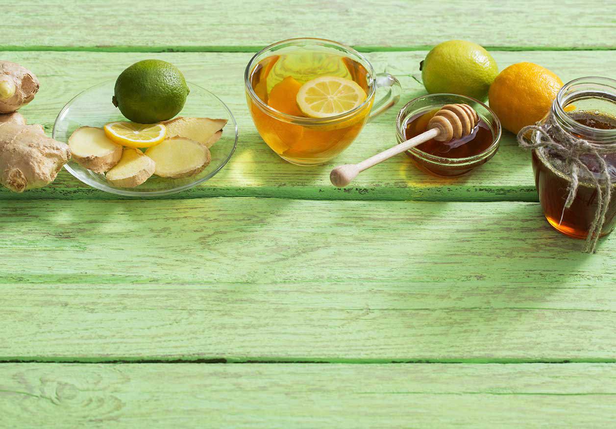 Eine Glas­tasse mit heißem Ingwertee und Zitronenscheiben steht auf einem grünen Holztisch, umgeben von frischem Ingwer, Ingwerscheiben auf einem Teller, ganzen Zitronen und Limetten, einer kleinen Schale mit Honig und Honiglöffel sowie einem mit Schnur gebundenen Teeglas.