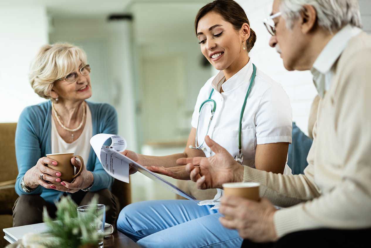O tânără asistentă medicală în haine de lucru albe, cu stetoscop, discută cu un cuplu în vârstă într-un mediu familial. Ea ține un document în mână și le explică conținutul cu un zâmbet prietenos. Femeia în vârstă ține o ceașcă de cafea în timp ce bărbatul în vârstă gesticulează. Scena transmite consiliere profesională și empatică pentru îngrijire într-o atmosferă relaxată.
