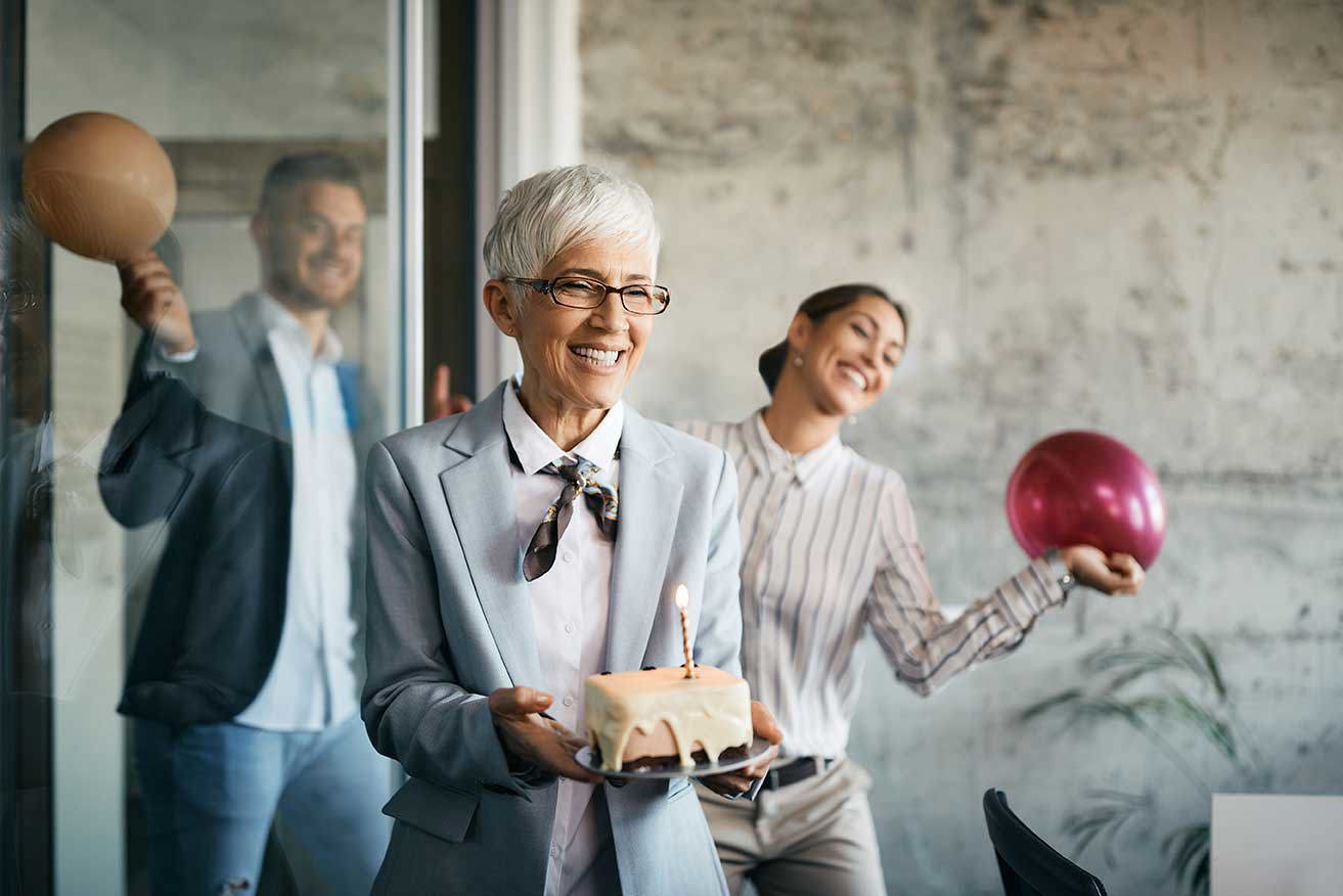 Eine Gruppe glücklicher Geschäftsleute, die in einem Büro eine Überraschungsparty feiern. Eine Person hält einen Geburtstagskuchen mit einer brennenden Kerze, während zwei andere Luftballons im Hintergrund halten.