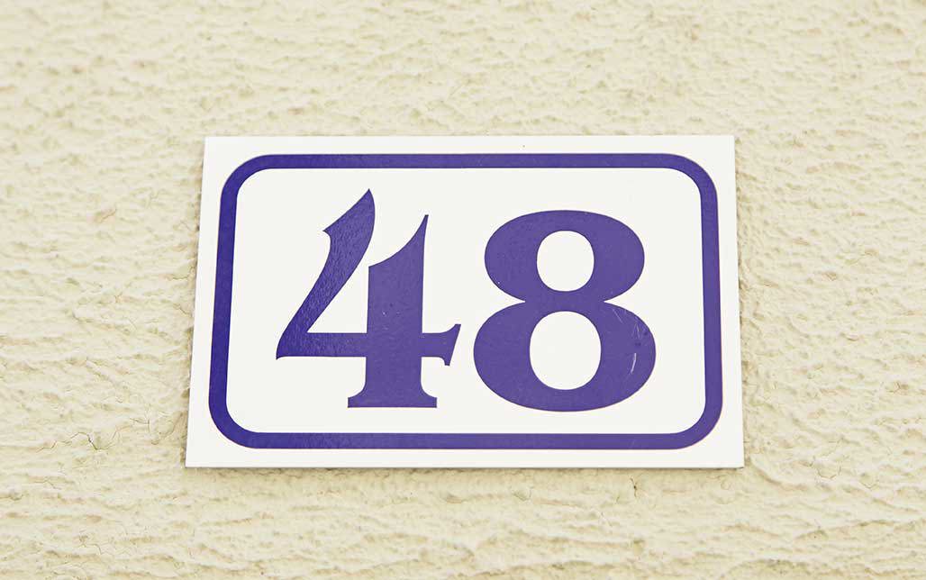 „Placă dreptunghiulară albă cu numărul casei 48 în culoare violet, montată pe un perete deschis la culoare, texturat.“