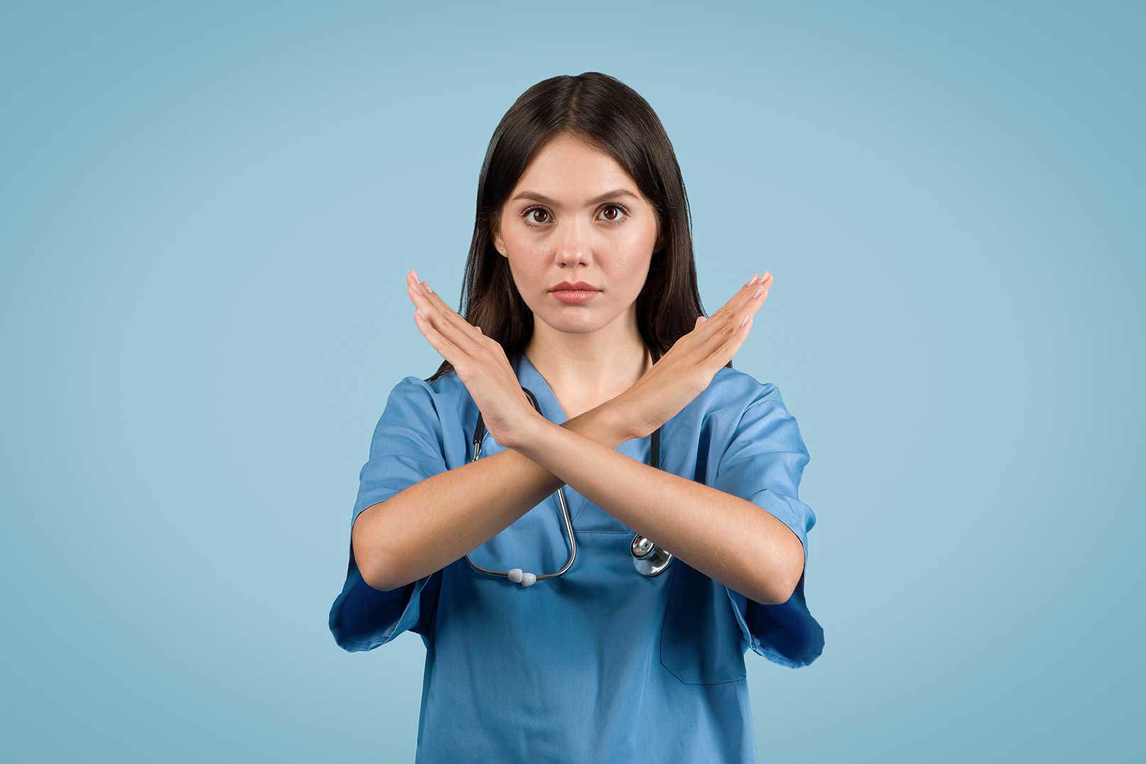 Junge Pflegekraft in blauer Uniform mit ernstem Gesichtsausdruck zeigt mit überkreuzten Armen ein Stoppsignal vor blauem Hintergrund.