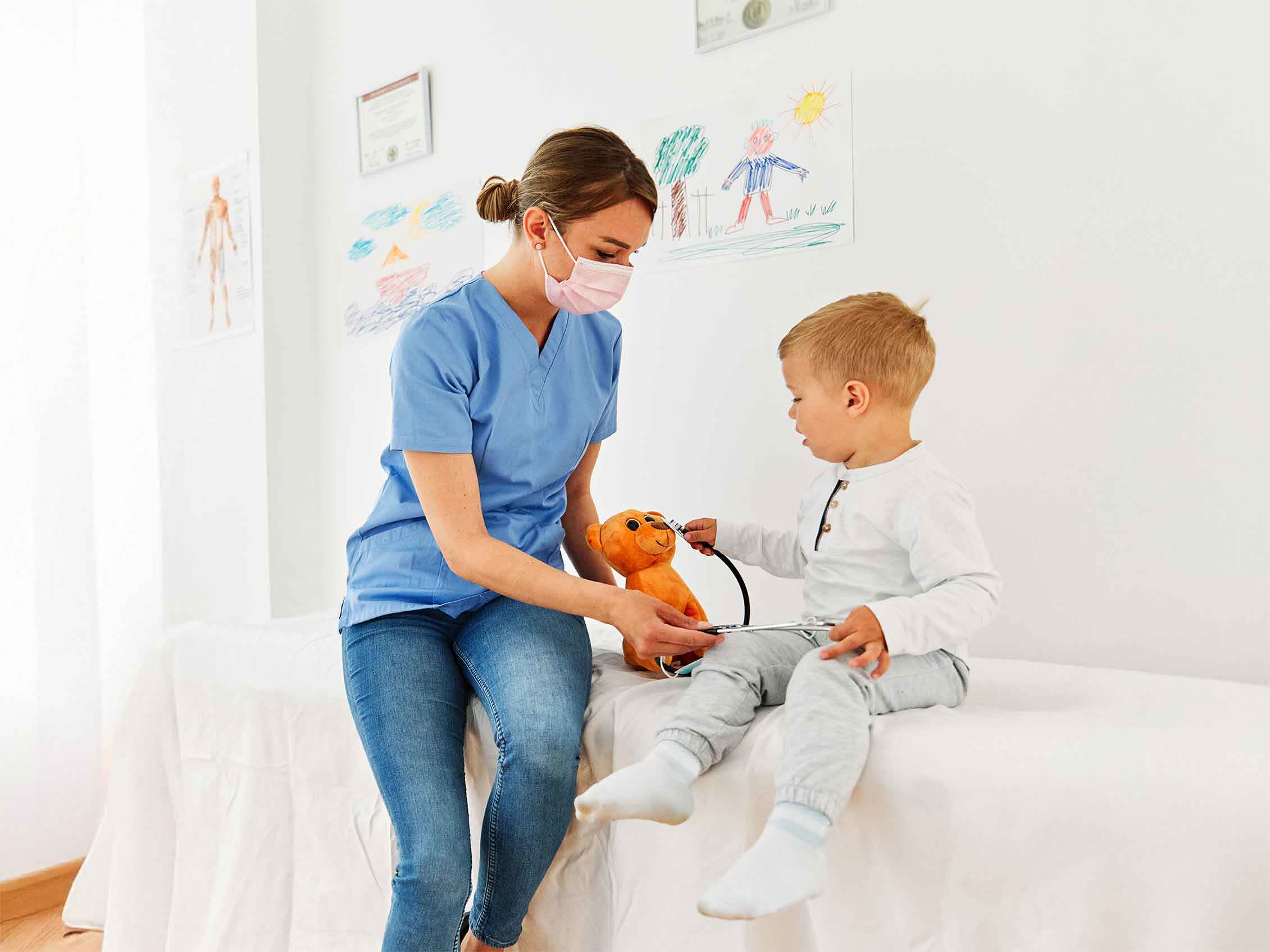 Krankenschwester in blauer Arbeitskleidung beruhigt einen kleinen Jungen während einer Untersuchung mit einem Spielzeug-Stethoskop und einem Stofftier