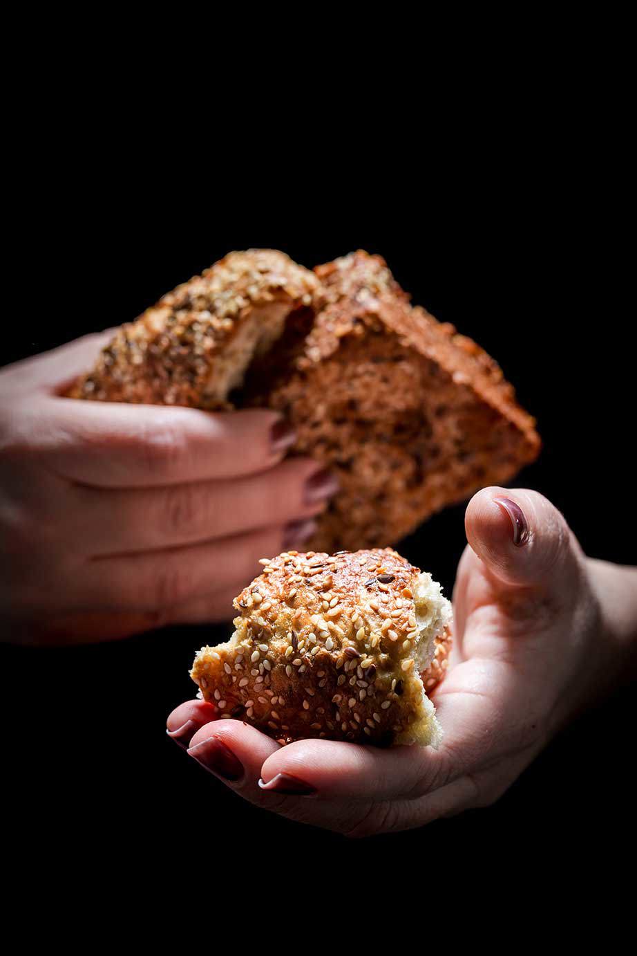 Hände halten Stücke frisches Brot mit Sesamsamen, vor dunklem Hintergrund.