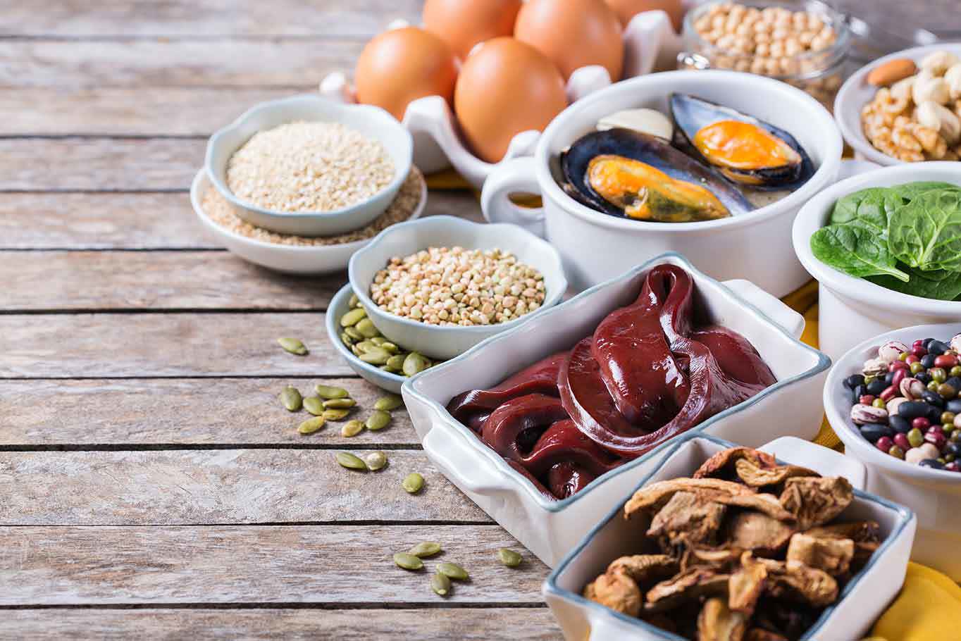 Eine Auswahl an eisenreichen Lebensmitteln, darunter Leber, Eier, Sesam, Kürbiskerne, Hülsenfrüchte und grünes Gemüse, auf einem Holztablett, die eine gesunde Ernährung fördern.