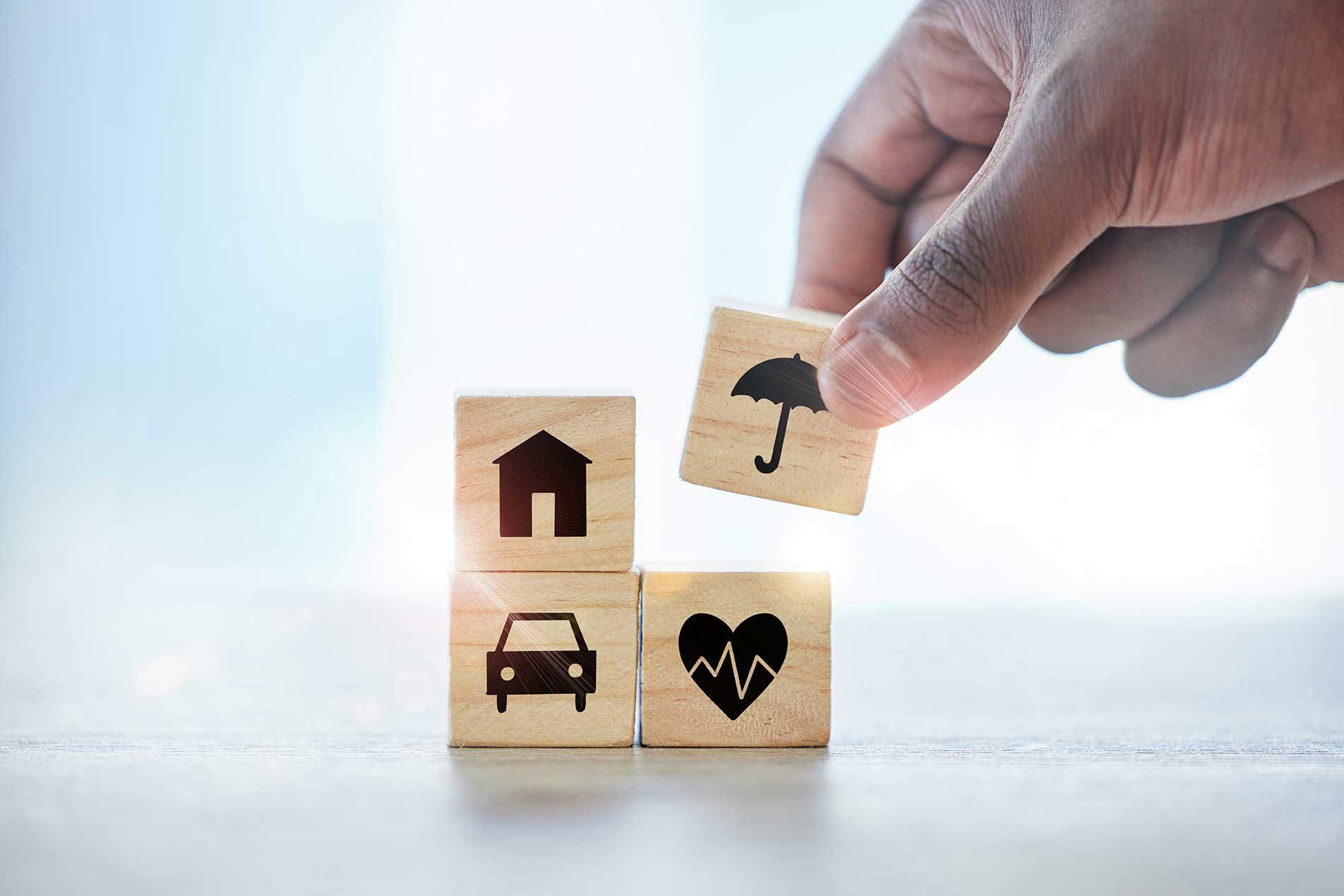 Eine Hand platziert einen Holzblock mit einem Regenschirm-Symbol auf einem Stapel von Blöcken. Die Blöcke sind mit Symbolen für ein Haus, ein Auto, ein Herzschlag und einen Regenschirm angeordnet und symbolisieren verschiedene Formen von Schutz und Sicherheit.