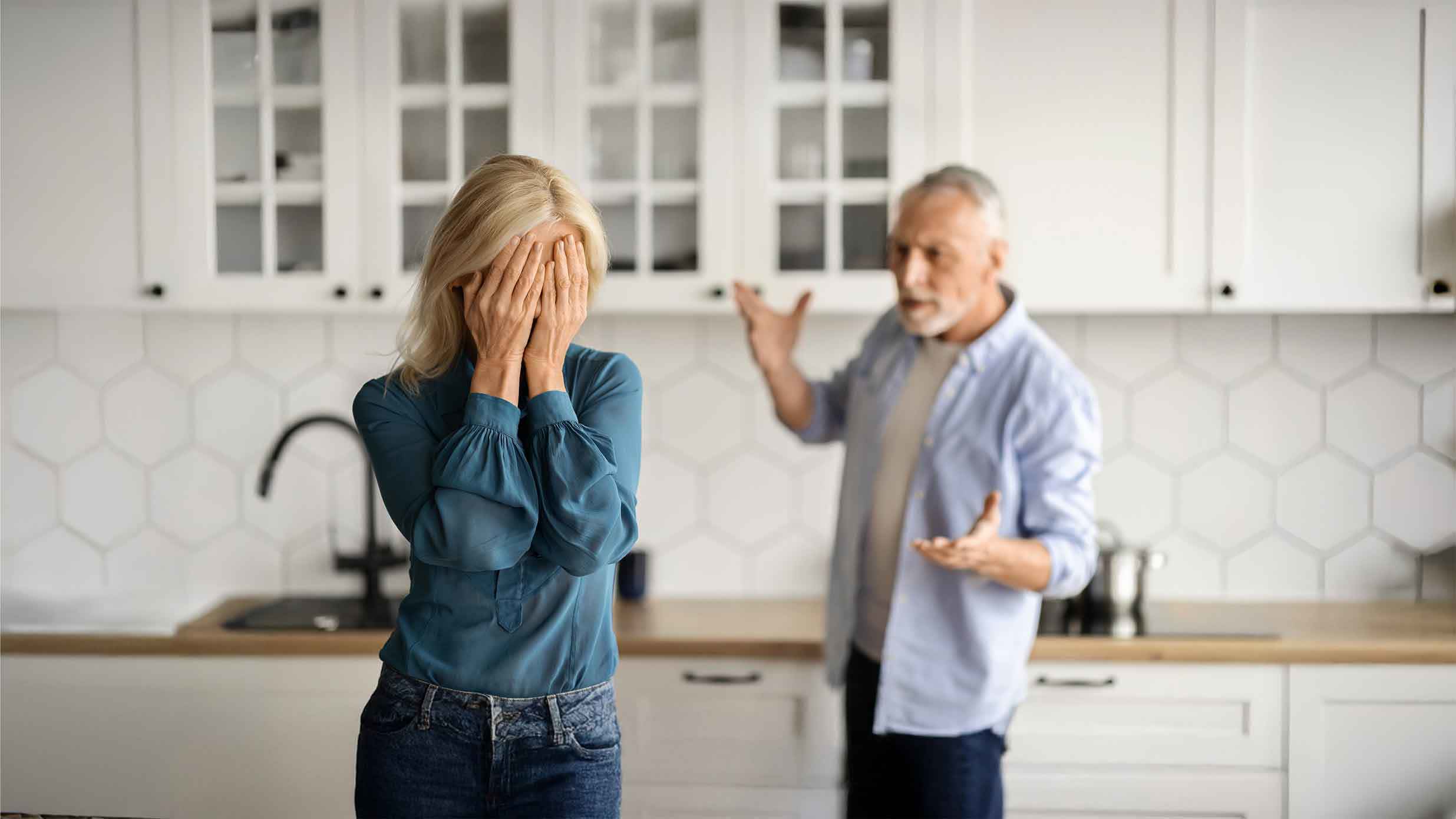 Eine ältere Frau in einer blauen Bluse, die ihr Gesicht aus Verzweiflung bedeckt, während ein älterer Mann in einem hellen Hemd im Hintergrund mit Frustration gestikuliert. Sie befinden sich in einer modernen Küche mit einem hexagonalen Fliesenspiegel.