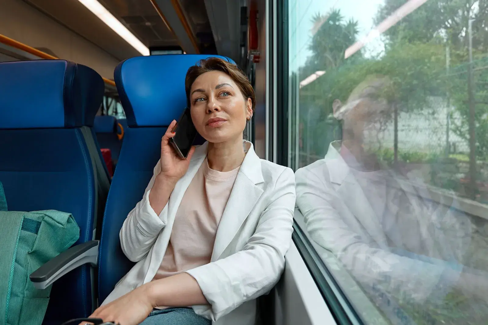 Frau sitzt im Zug am Fenster und telefoniert mit dem Smartphone, während die Landschaft vorbeizieht.