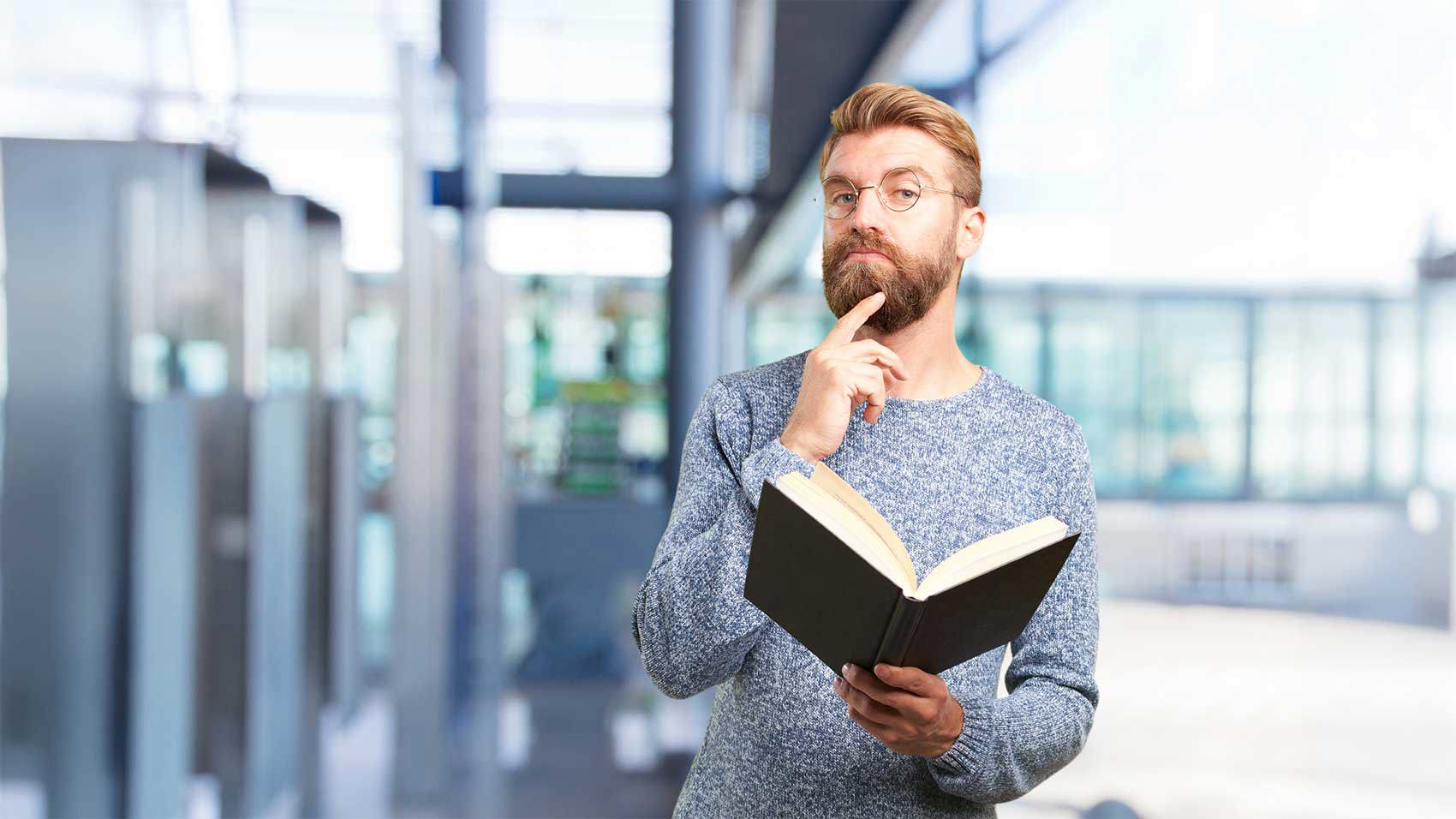 Ein bärtiger Mann mit Brille und blauem Pullover steht in einem Innenraum, hält ein geöffnetes Buch und berührt nachdenklich sein Kinn.