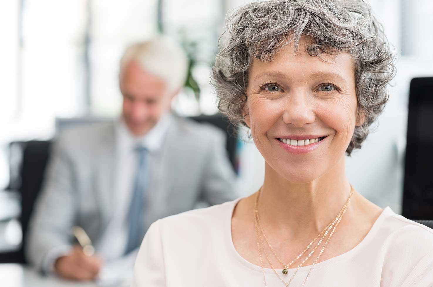 Ein Porträt einer lächelnden älteren Geschäftsfrau in einer hellen Bluse, mit einem unscharfen männlichen Kollegen im Hintergrund. Sie wirkt selbstbewusst und glücklich.