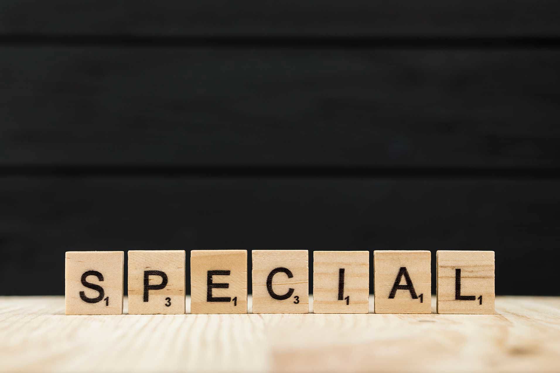 Das Wort „SPECIAL“ ist mit hölzernen Scrabble-Buchstaben auf einer hellen Holzoberfläche in einer geraden Linie gelegt, vor einem dunklen, unscharfen Hintergrund.