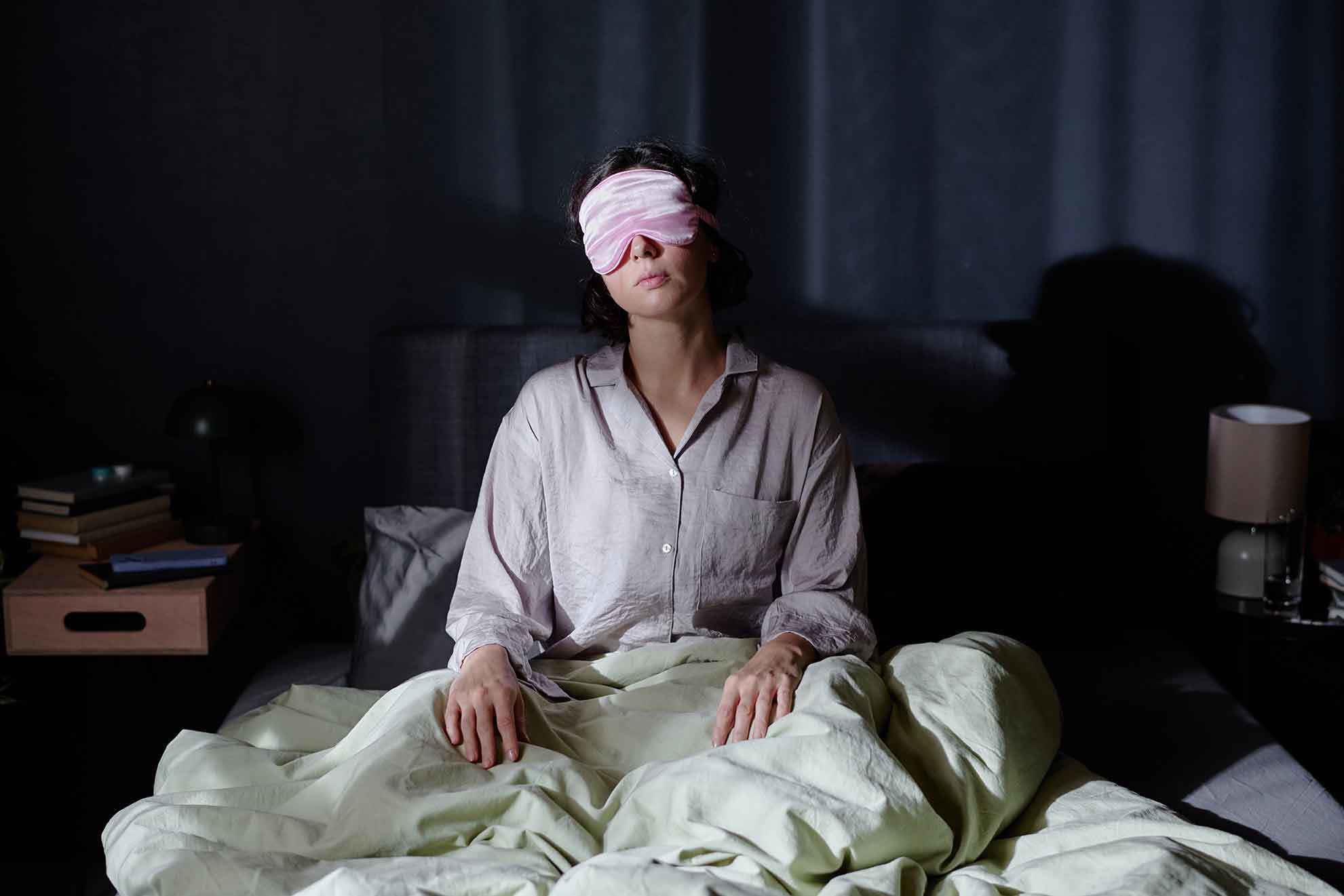 „Frau sitzt nachts wach im Bett und trägt eine Schlafmaske; gedämpftes Licht in einem dunklen Schlafzimmer.“