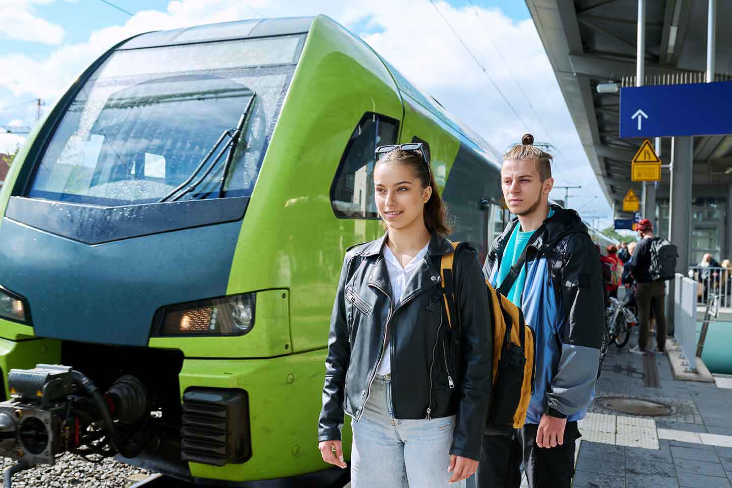 Junge Frau und junger Mann mit Rucksäcken warten am Bahnhof vor einem grünen Zug