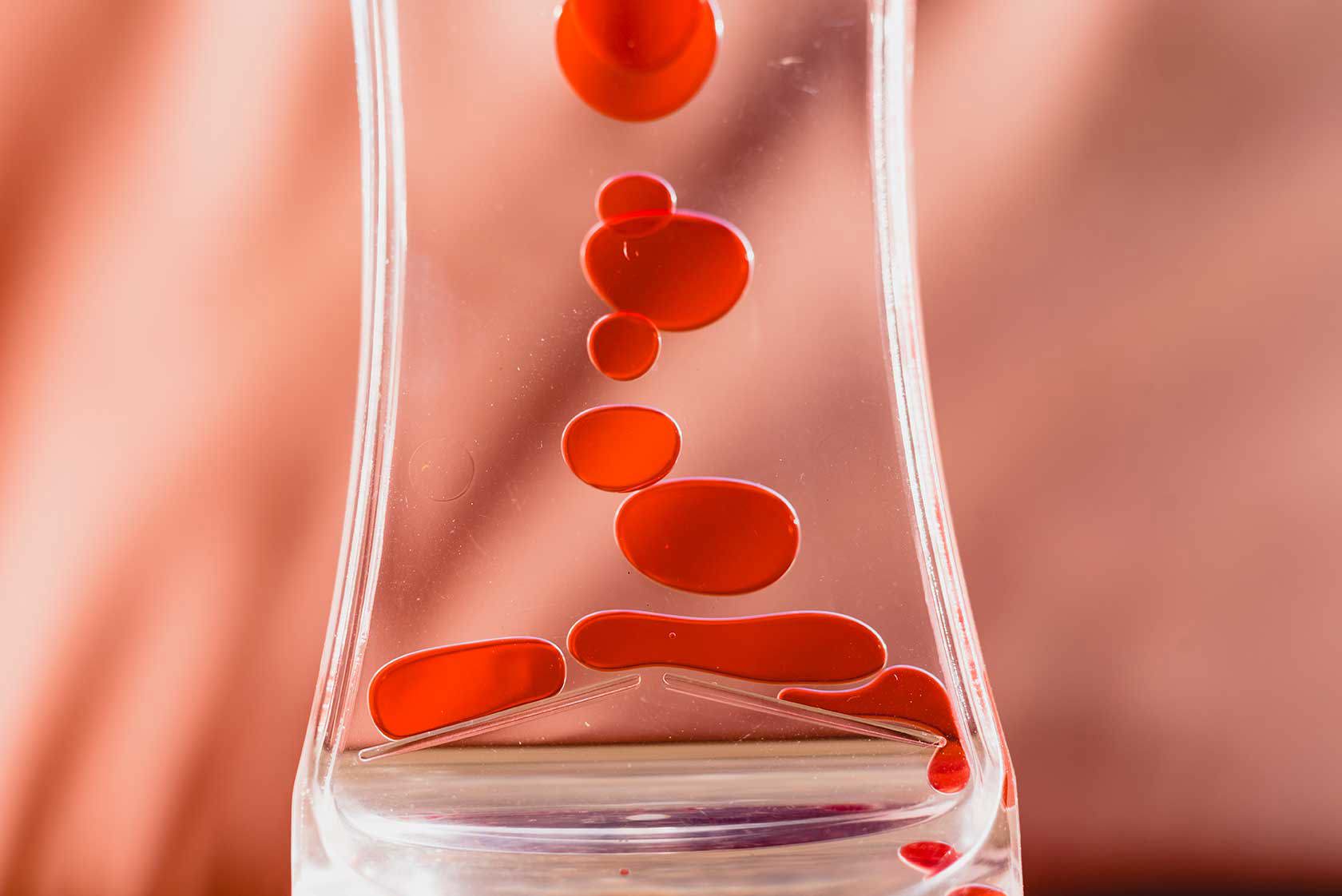 Nahaufnahme roter Tropfen, die in einer durchsichtigen Flüssigkeit fallen und an Blut erinnern, vor einem unscharfen rosafarbenen Hintergrund.