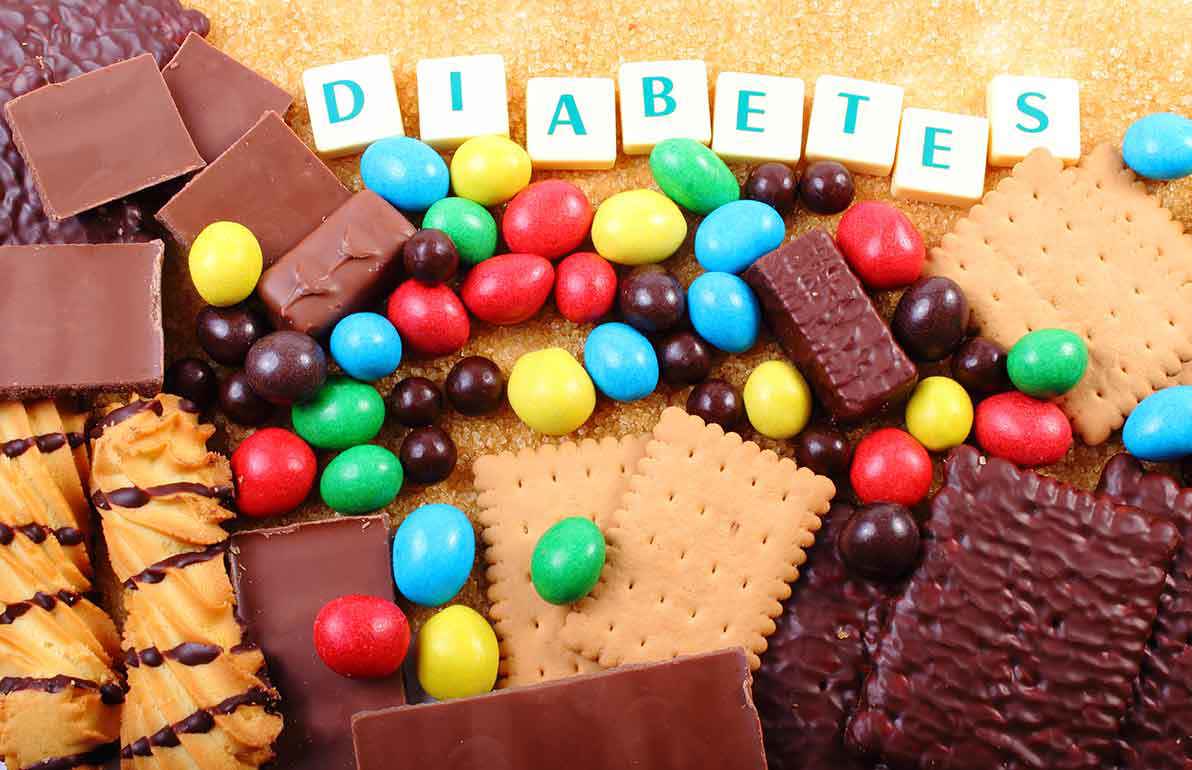 Bunte Bonbons, Schokoladenstücke und Kekse, die auf braunem Zucker arrangiert sind, mit Buchstabensteinen, die oben das Wort „DIABETES“ bilden.
