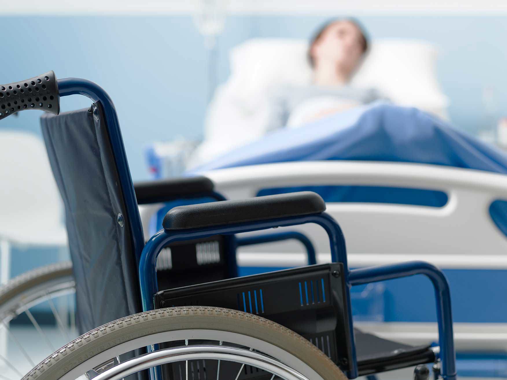 Ein Rollstuhl steht im Vordergrund eines Krankenzimmers, während im Hintergrund unscharf eine Person in einem Krankenhausbett liegt.