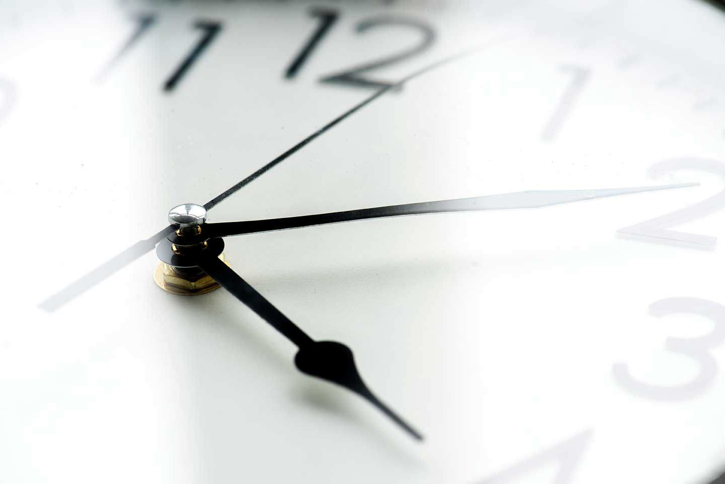 Nahaufnahme der Zeiger einer Uhr auf einem weißen Zifferblatt mit unscharfen Zahlen im Hintergrund.