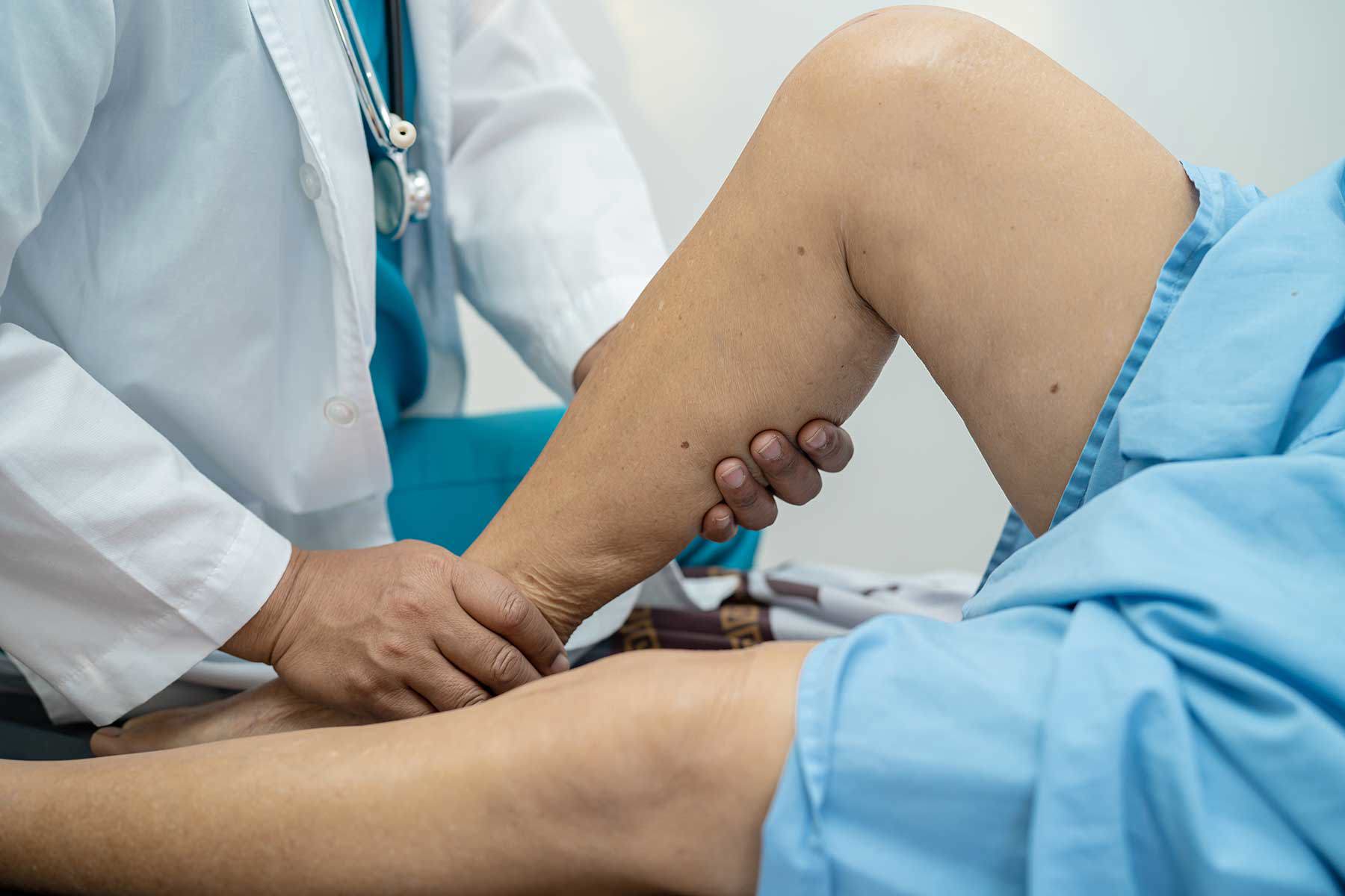 Nahaufnahme eines asiatischen Arztes oder Physiotherapeuten, der das Bein eines Patienten in einer klinischen Umgebung untersucht und massiert. Der Patient liegt auf einem Behandlungstisch, während der Therapeut das Knie des Patienten hält und bewegt.