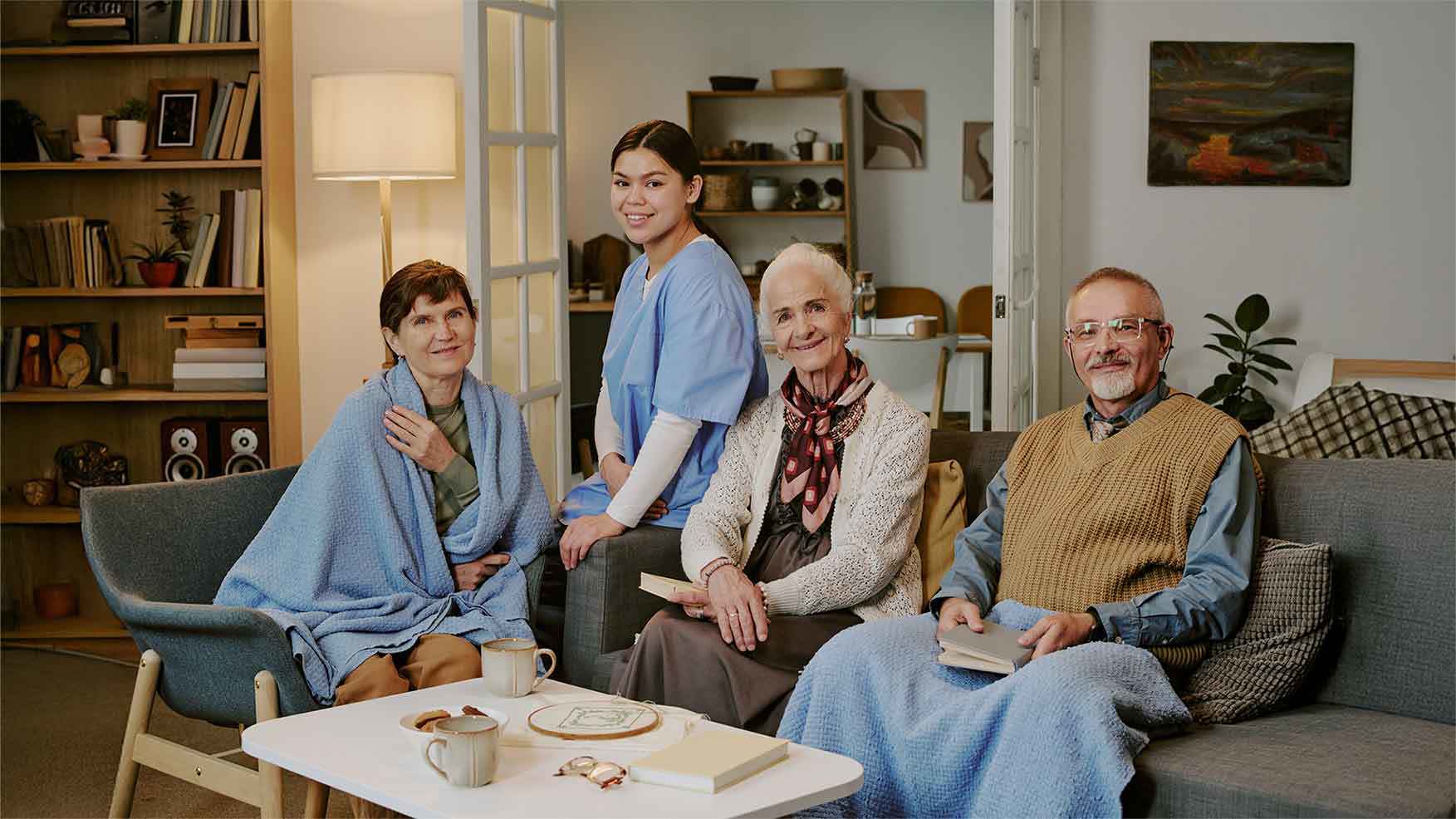 Eine lächelnde Pflegekraft in blauer Arbeitskleidung steht zwischen drei älteren Menschen, die auf einem Sofa in einem gemütlichen Wohnzimmer sitzen und Bücher sowie Tassen mit Tee halten.