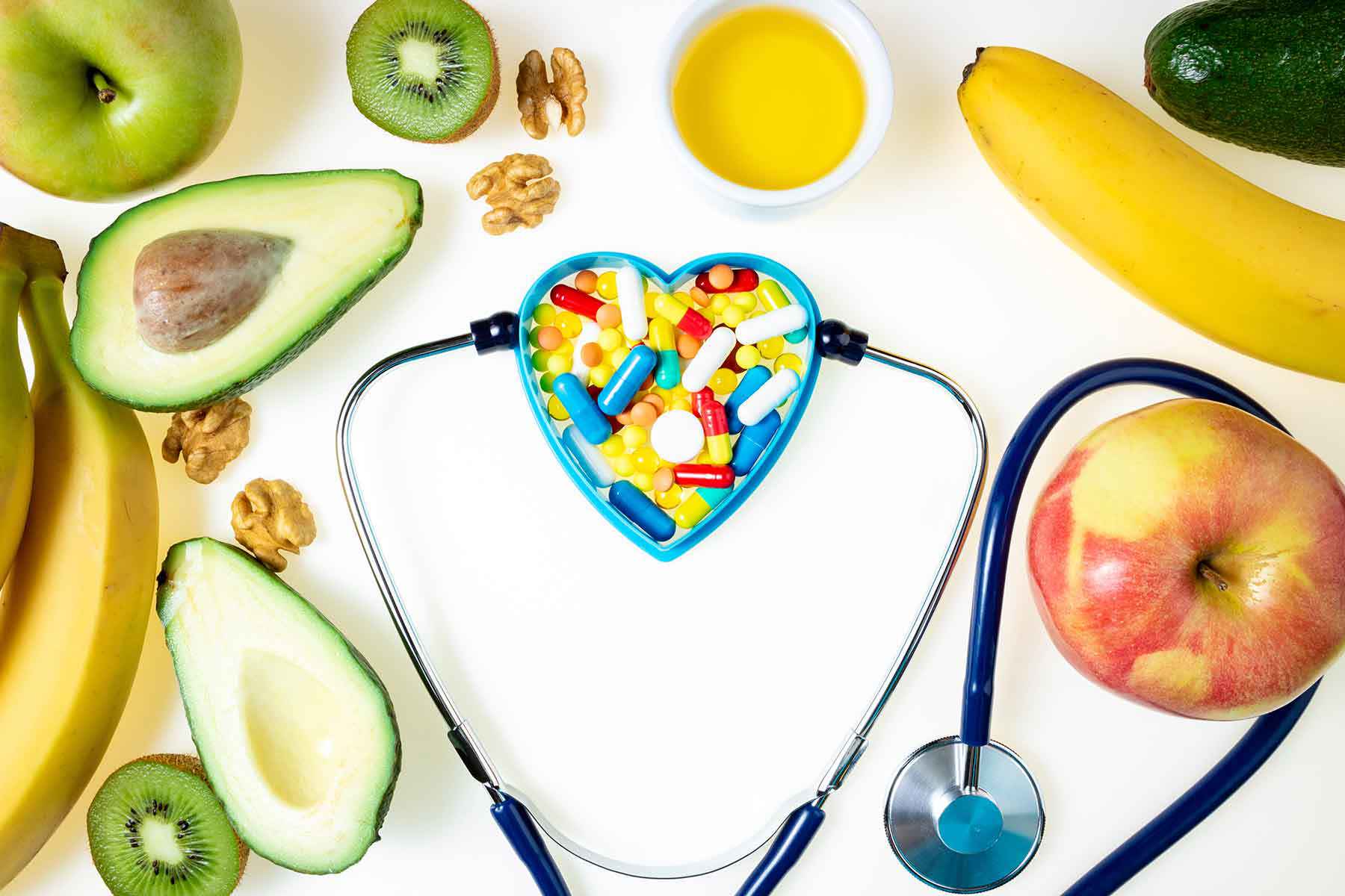 Ein Stethoskop umrahmt ein herzförmiges Schälchen voller bunter Vitaminkapseln, umgeben von gesunden Lebensmitteln wie Avocado, Kiwi, Banane und Apfel – Symbol für die Verbindung zwischen Ernährung und Herzgesundheit.