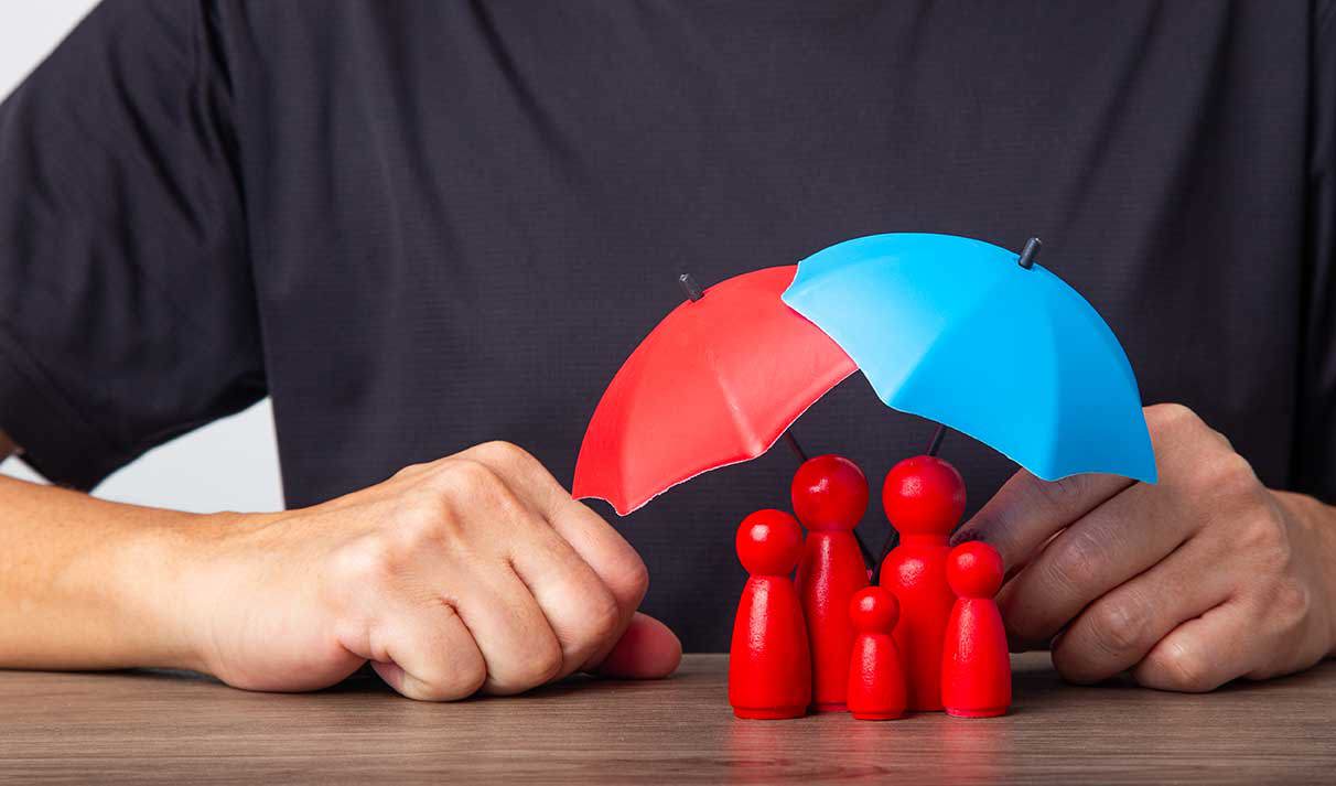 Eine Hand hält zwei bunte Regenschirme, einen roten und einen blauen, über einer kleinen Gruppe von roten Figuren, die eine Familie darstellen. Die Figuren sind unter den Regenschirmen geschützt, was Schutz und Fürsorge symbolisiert.