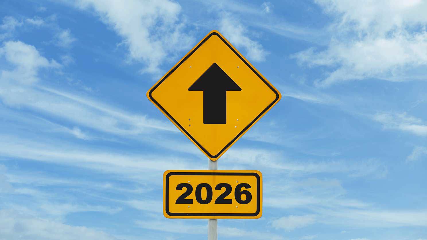Gelbes Verkehrsschild mit schwarzem Pfeil nach oben, darunter ein Zusatzschild mit der Aufschrift „2026“, vor blauem Himmel mit Wolken.