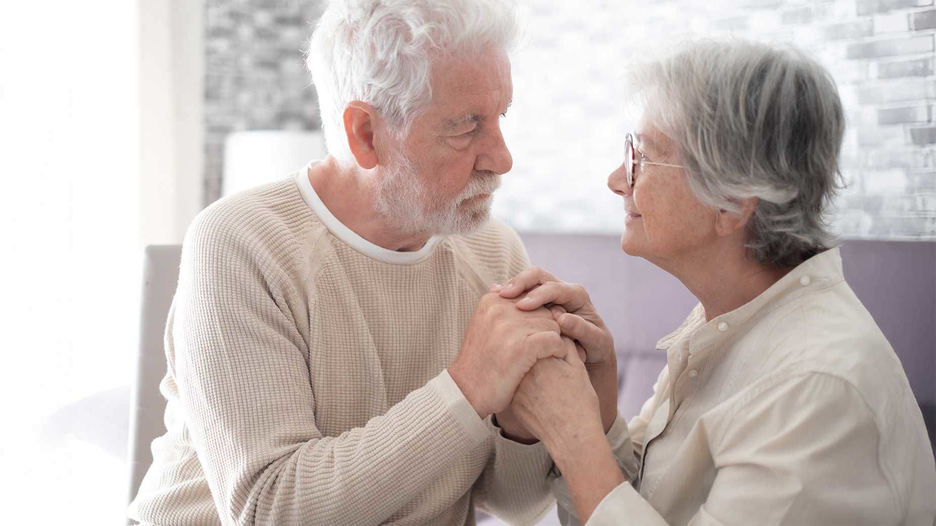 Ein älteres Paar sitzt sich in einem hellen Wohnzimmer gegenüber und hält sich liebevoll an den Händen, während sie sich ruhig und aufmerksam in die Augen schauen.