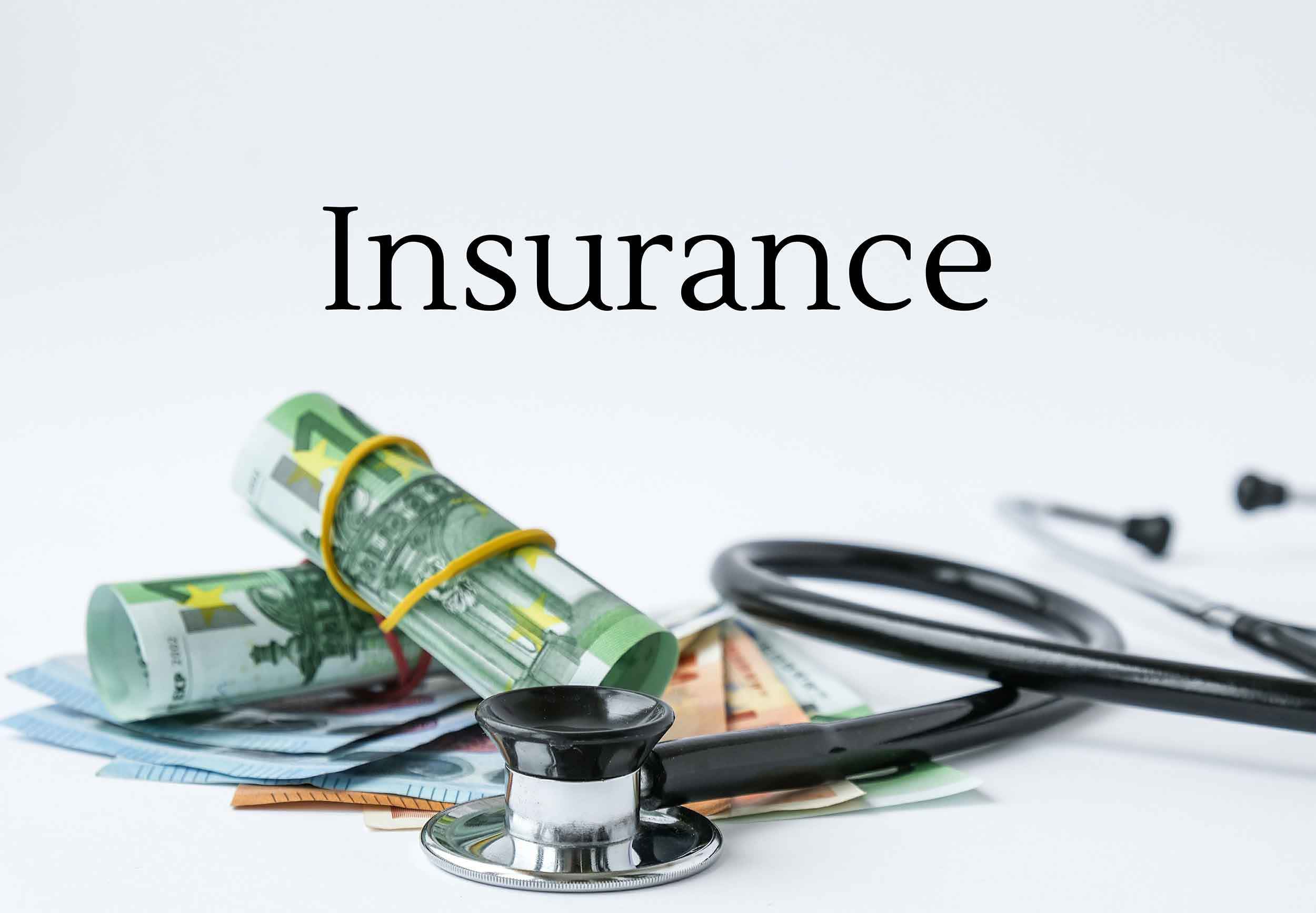 Ein Stapel Geldscheine, darunter Euro, mit einem Gummiband zusammengehalten, liegt neben einem Stethoskop. Das Wort „Insurance“ (Versicherung) steht prominent im Hintergrund.
