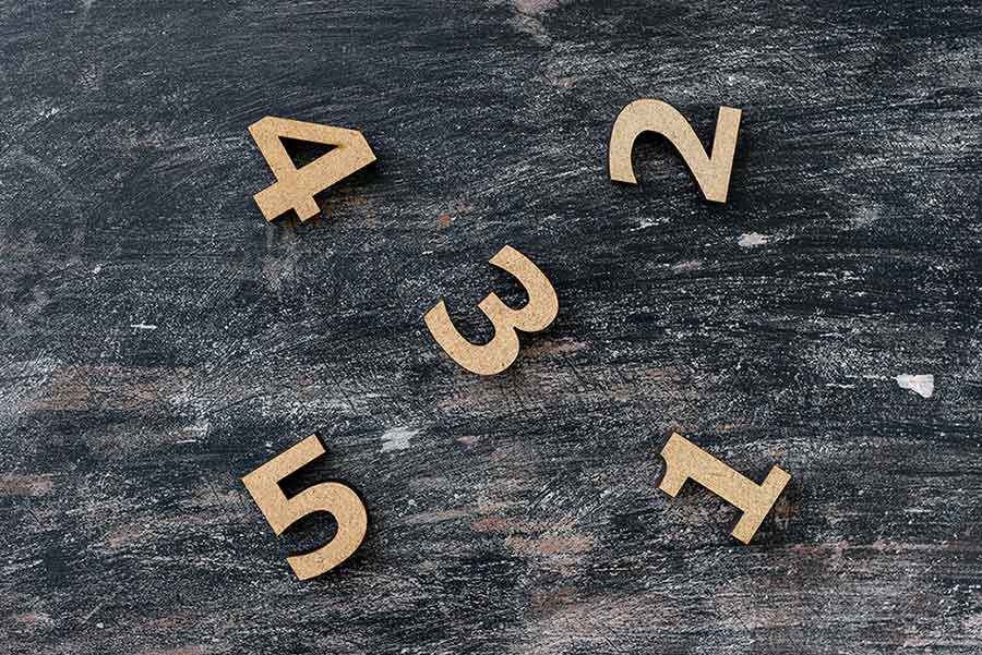 Holzzahlen von 1 bis 5 liegen verstreut auf einer dunklen, verwitterten Holzoberfläche, mit Abstand zueinander und aus der Vogelperspektive aufgenommen.