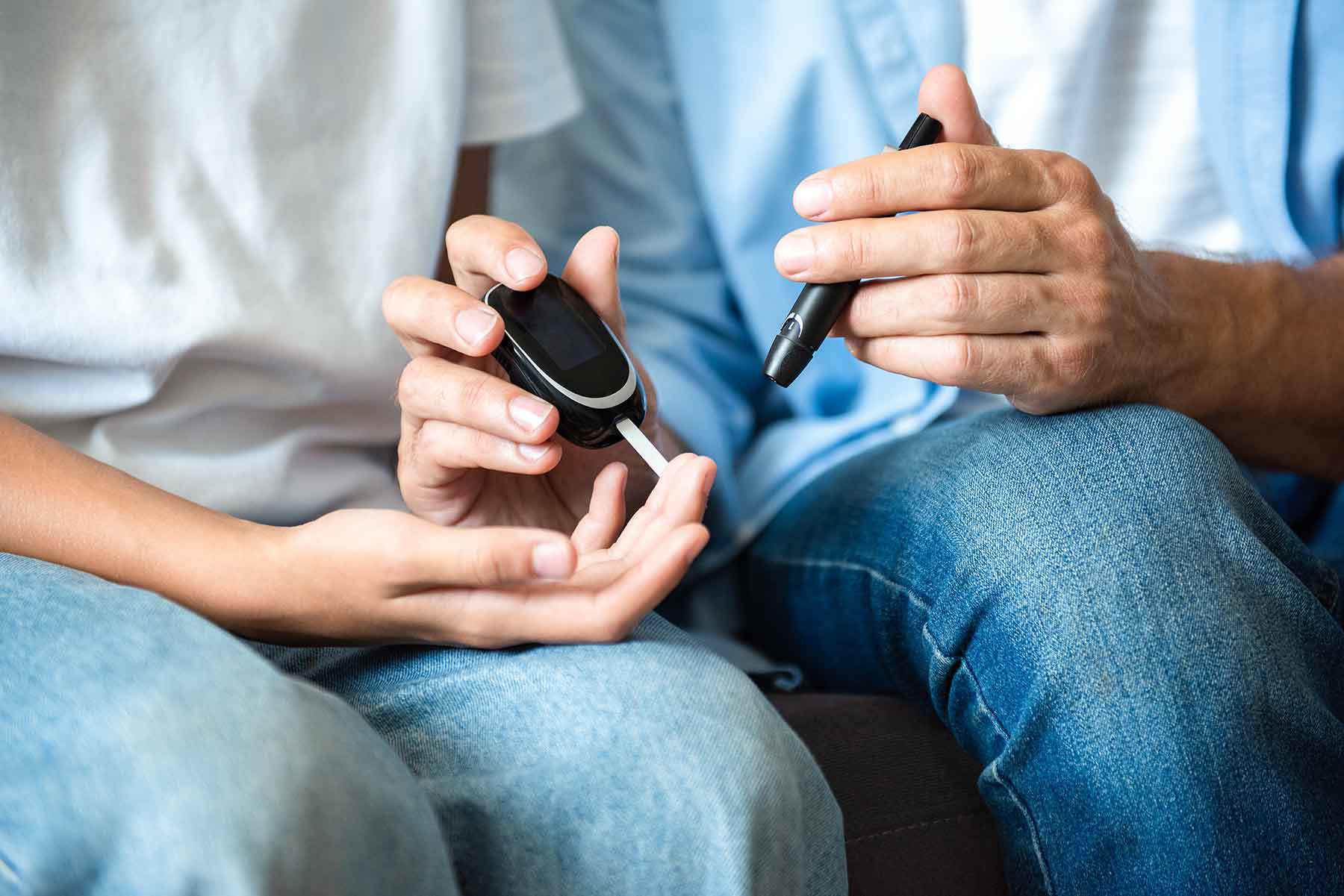 Nahaufnahme eines Erwachsenen, der mit einem Glukometer und einer Stechhilfe den Blutzuckerspiegel eines Kindes misst, indem er in dessen Finger sticht, während sie zusammen auf einem Sofa sitzen.