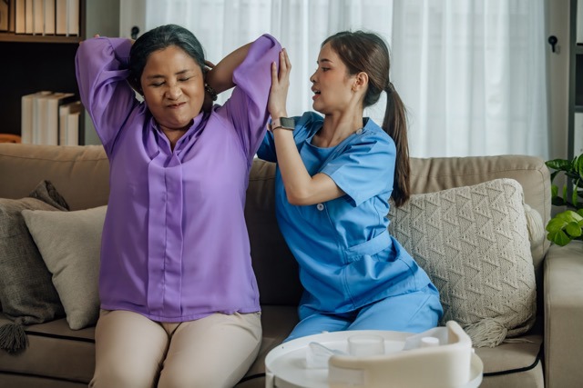 Betreuerin in blauer Dienstkleidung hilft einer älteren Frau in lila Bluse bei Armübungen auf einem Sofa und unterstützt sie dabei, ihre Arme hinter den Kopf zu strecken, in einer unterstützenden häuslichen Umgebung.