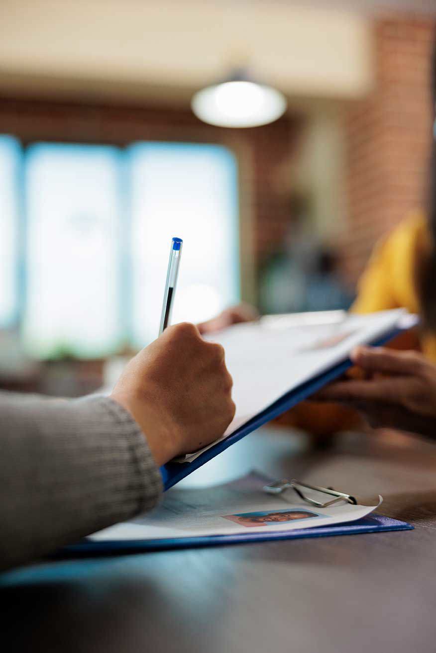 Eine Nahaufnahme der Hand einer Frau, die einen Stift hält und Dokumente auf einem Klemmbrett unterschreibt. Im Hintergrund ist die Hand einer anderen Person zu sehen, was auf eine geschäftliche oder professionelle Umgebung hindeutet. Ein Paar Brillen ist ebenfalls neben den Papieren sichtbar.