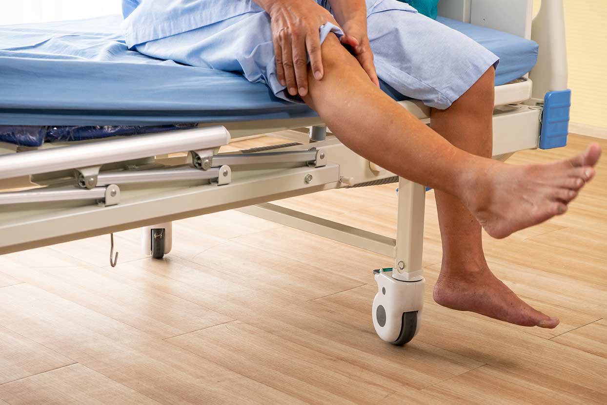 Ein älterer asiatischer Mann in einem Krankenhaushemd sitzt auf einem Krankenhausbett und hält sich schmerzerfüllt das Knie. Der Raum ist mit einem Krankenhausbett und einem Holzboden ausgestattet.