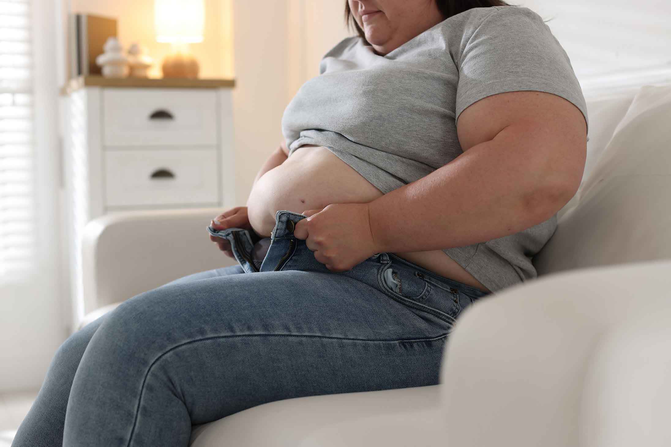 “Overweight person sitting on a sofa and trying to close tight jeans.”