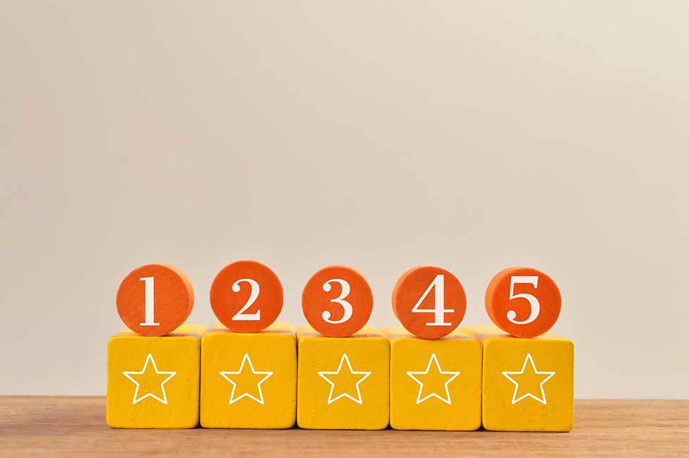 Fünf orangefarbene runde Plättchen mit den Zahlen 1 bis 5 stehen auf gelben Holzklötzen mit weißen Sternsymbolen, in einer Reihe auf einer Holzoberfläche vor einem schlichten Hintergrund angeordnet.