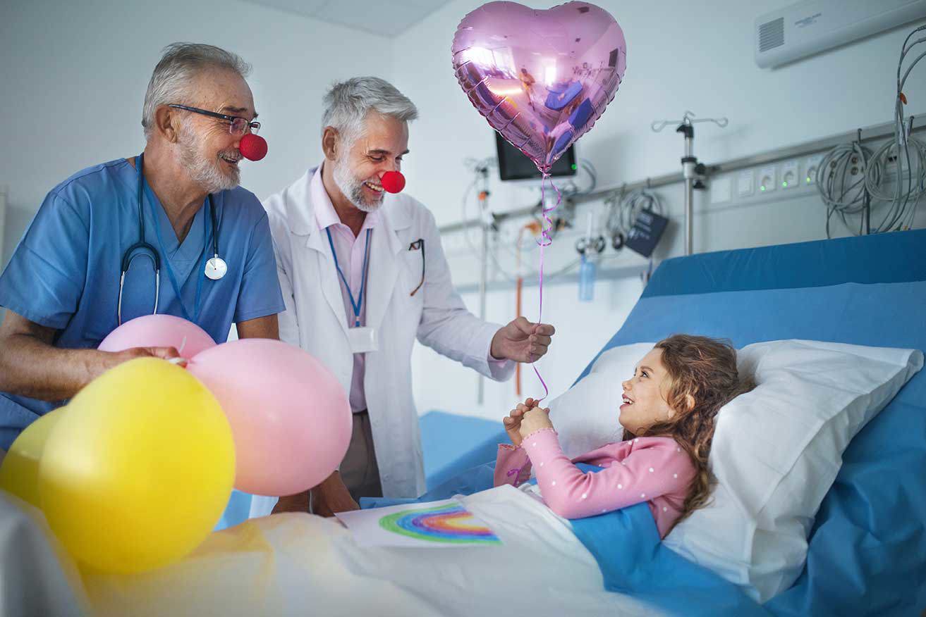 Zwei Ärzte mit roten Clownnasen bringen einem jungen Patienten im Krankenhausbett Freude, indem sie Ballons und einen herzförmigen Ballon überreichen, um ihren Geburtstag mit einer bunten und fröhlichen Atmosphäre zu feiern.
