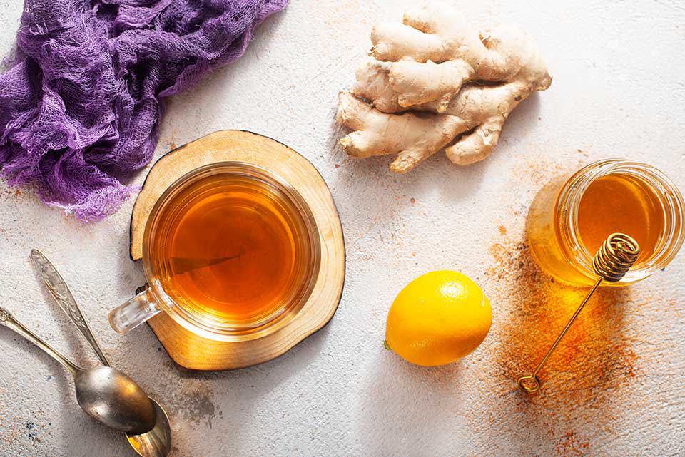 Draufsicht auf eine Glastasse mit bernsteinfarbenem Ingwertee auf einem Holzuntersetzer, daneben frische Ingwerwurzel, eine ganze Zitrone, ein Glas Honig mit Honigquirl, zwei Löffel und ein violettes Tuch auf einer hellen, strukturierten Oberfläche mit verstreutem Gewürzpulver.