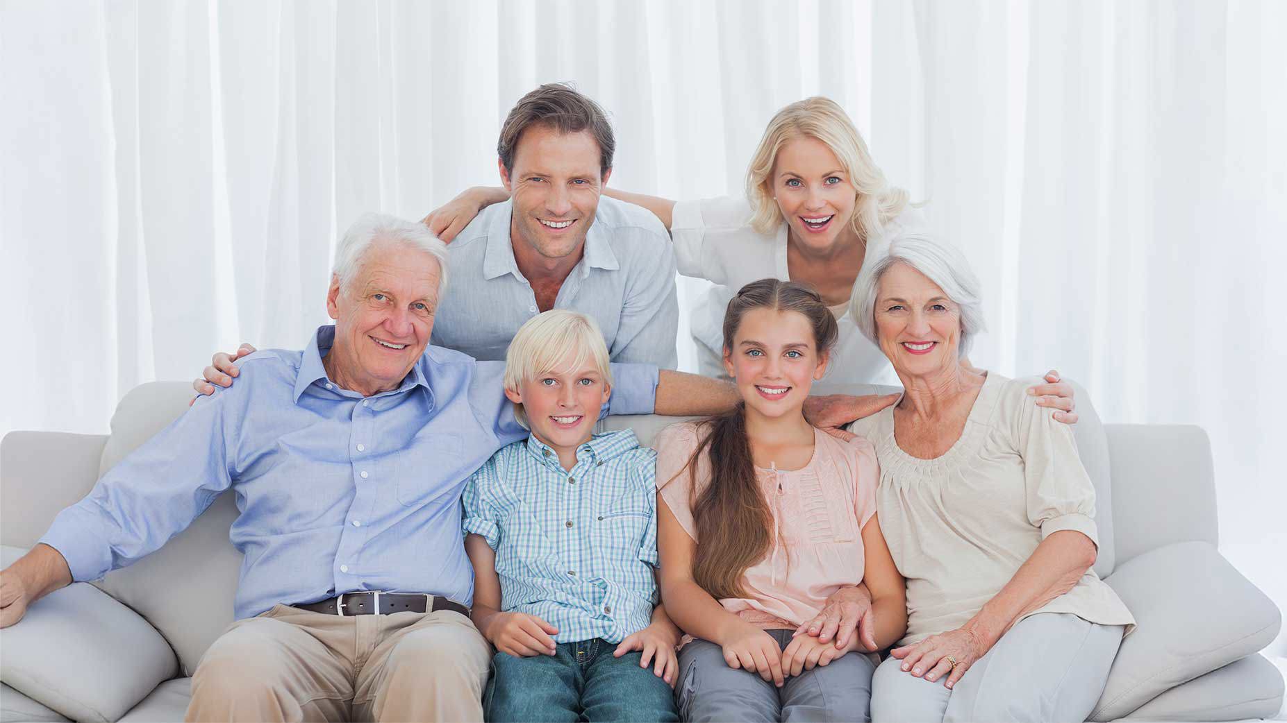 Eine glückliche Mehrgenerationenfamilie sitzt gemeinsam auf einem Sofa und lächelt in die Kamera – Großeltern, Eltern und zwei Kinder in einem hellen Wohnzimmer.