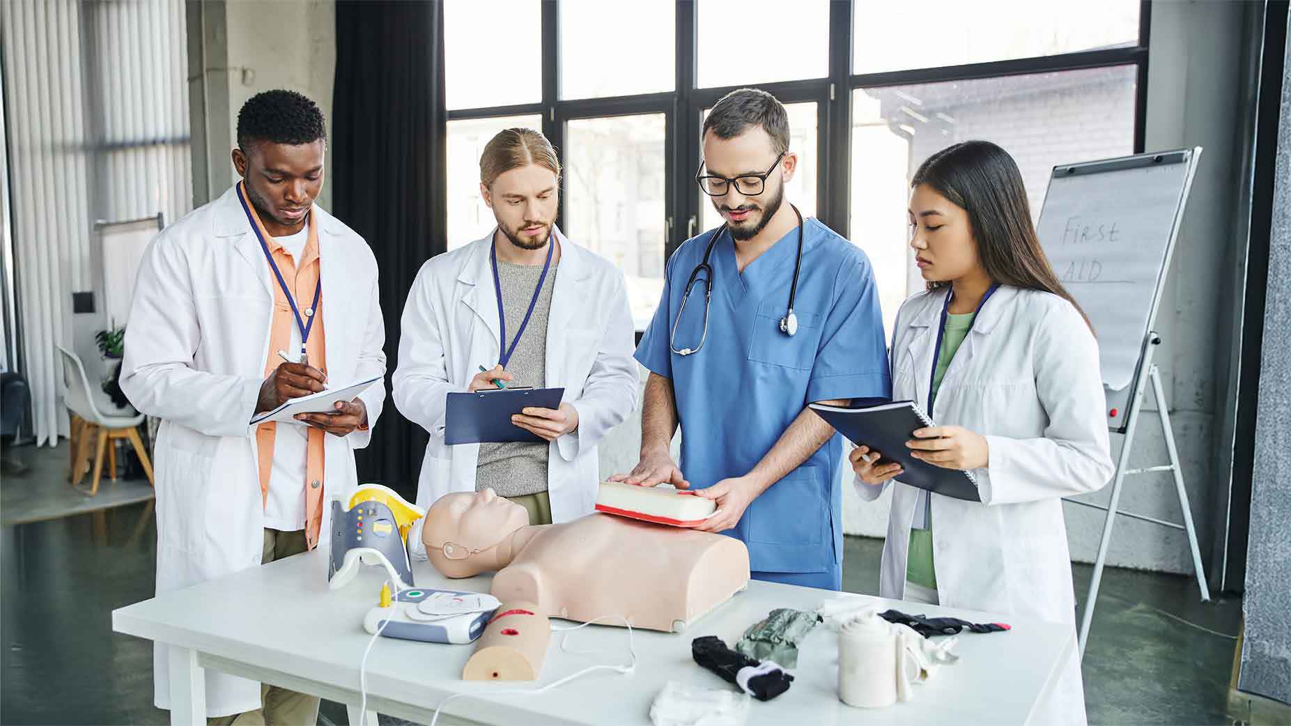 Eine Gruppe von medizinischen Auszubildenden in Laborkitteln und OP-Kleidung übt Erste Hilfe und Wiederbelebung an einer Übungspuppe, verwendet einen Defibrillator und macht sich während einer Schulung Notizen.