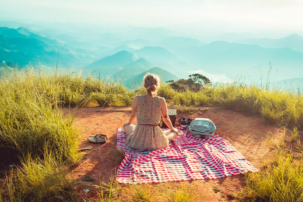 Een vrouw die op een picknick-kleed zit in de zon en groene heuvels en bergen in de verte bekijkt.