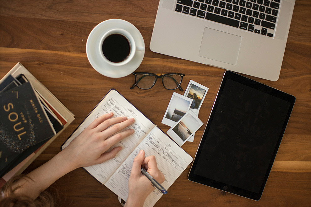 A person works on their travel journal next to photos, a pile of books, a tablet, a laptop, and a pair of glasses.