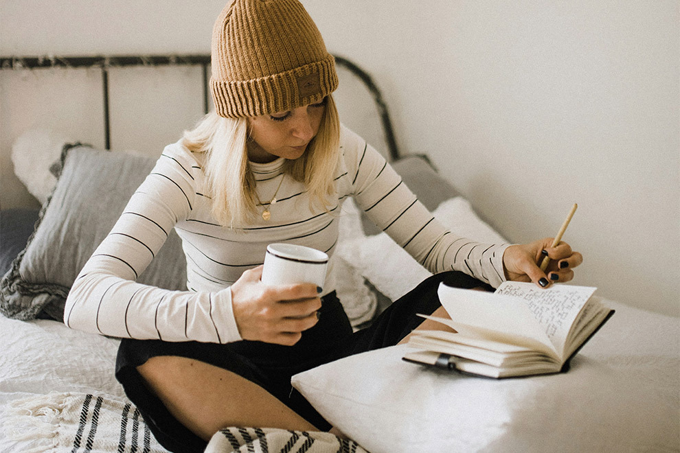 A woman sits in bed writing in her diary with a cup of coffee or tea in hand.