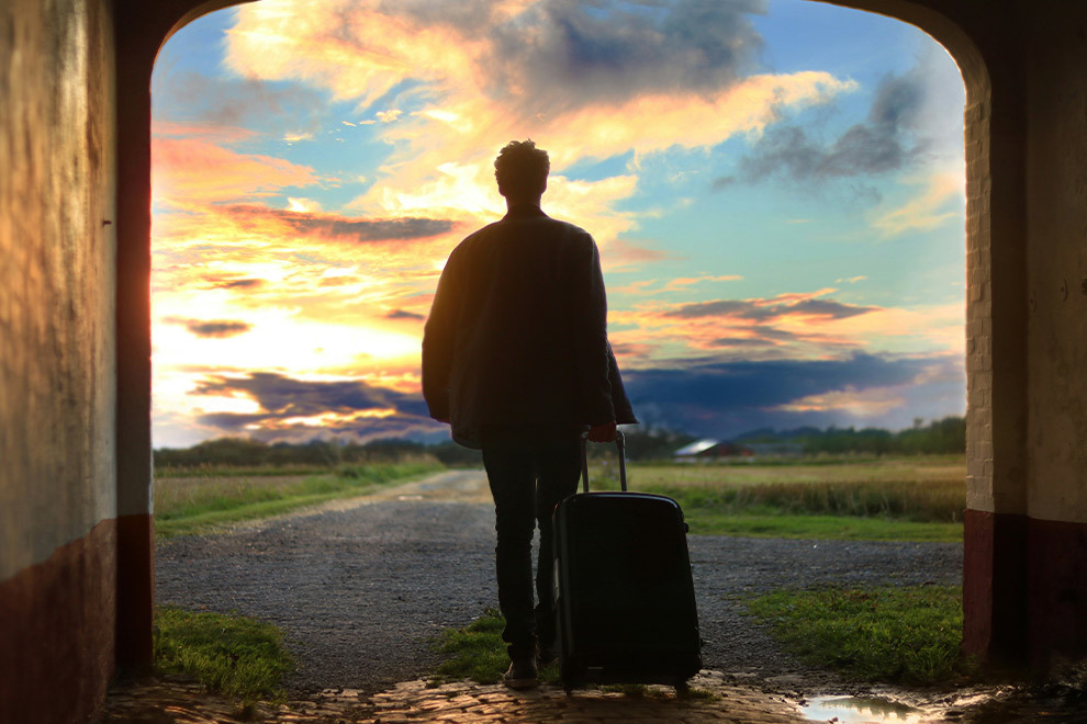 Een persoon loopt door een poort heen richting een veld tijdens de zonsopgang met zijn of haar koffer in hand.