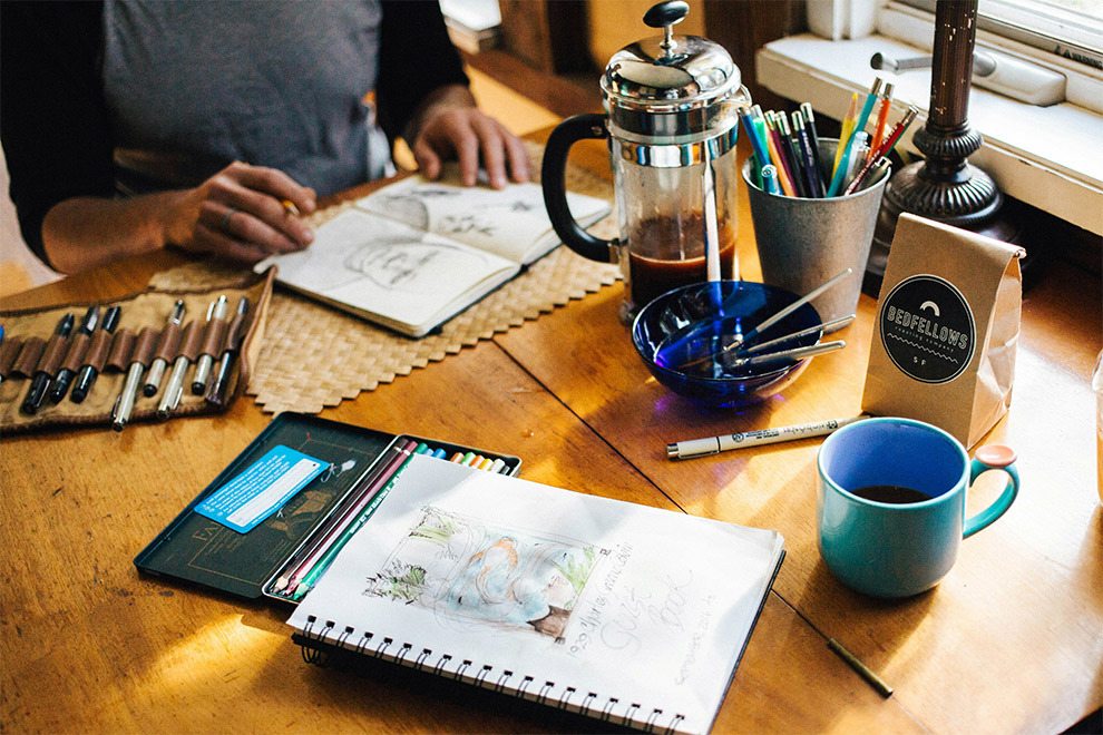 A person sits at a table with art supplies, working on a new piece on paper.