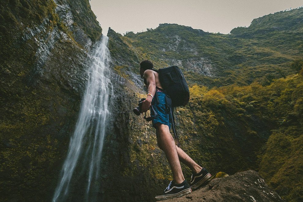 A man stands at the bottom of a waterfall with a camera in hand.