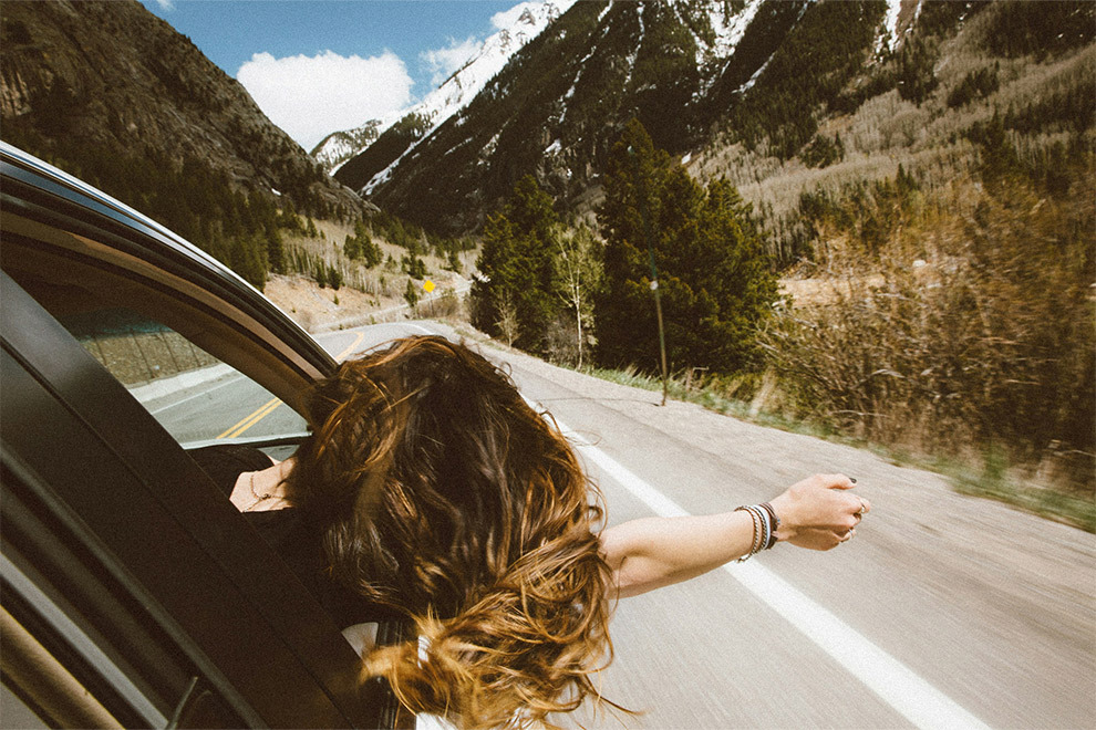 A woman hangs her arm and head out of a car's passenger seat window while driving down a wooded, mountainous road.